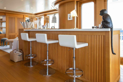 Three white bar stools stand before a wooden bar with a monkey statue on the right, while the background reveals a well-lit lounge area with large windows.