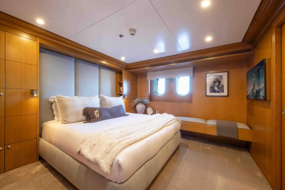A luxurious yacht bedroom features a plush king-sized bed in the foreground and a seating area with porthole windows in the background.