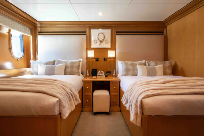 Two neatly made beds with plush throws flank a wooden nightstand with a stool in front, all set against a backdrop of warm wood paneling and upholstered headboards.