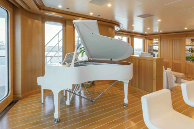 A gleaming white grand piano sits on a polished wooden floor in the foreground, while the background reveals a luxurious lounge area with large windows overlooking the water.