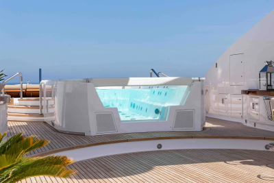 A modern, illuminated hot tub with clear sides sits on a wooden deck, with a bright blue sky and a distant structure in the background.