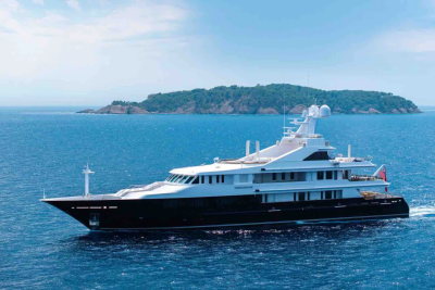 A sleek, dark-hulled yacht cruises on calm blue water with a lush, green island in the background under a hazy sky.
