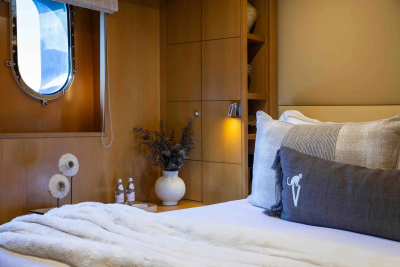 A cozy bedroom with a plush white blanket and decorative pillows in the foreground, and a round porthole window and wooden cabinetry in the background.