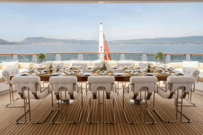 A long dining table set for a meal on a yacht's deck is surrounded by white chairs, with a calm sea and distant coastline visible in the background.