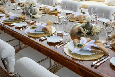 A long, polished wooden table is set for a formal meal with white chairs and elegant place settings in the foreground, while a blurred background suggests an outdoor setting with plush seating.