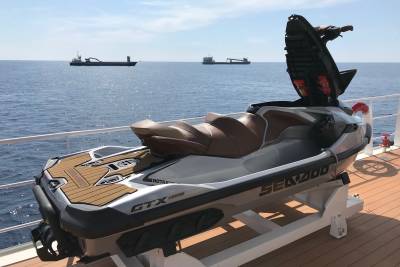 A silver and brown jet ski rests on a deck in the foreground, with two large ships sailing on a calm blue sea under a bright sky in the background.