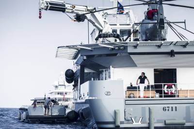 A man in white pants stands on the deck of a large yacht in the foreground, while a helicopter is visible on the upper deck of another vessel in the background.