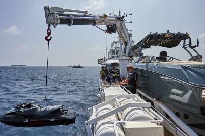 A jet ski is being lowered by a crane from a boat in the foreground, with a luxury yacht and a hazy horizon in the background.