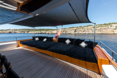 A spacious yacht deck with plush black loungers and pillows in the foreground is anchored near a rocky, sun-drenched coastline under a clear blue sky.