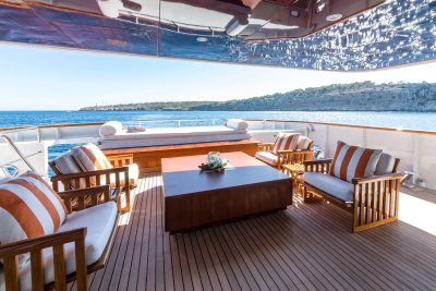 Wooden deck chairs and a coffee table are arranged on a yacht's deck, with a calm blue sea and a tree-covered coastline in the background.