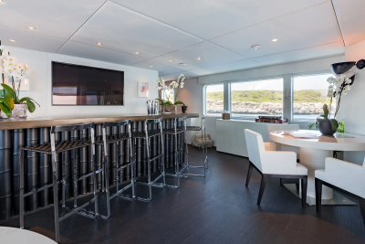 A modern yacht lounge features a bar with stools in the foreground and a dining area with a view of the ocean in the background.