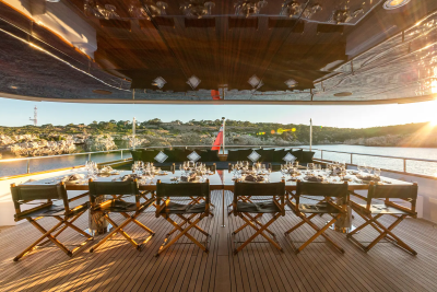A long dining table is set for a meal on a yacht deck, with a rocky coastline and the setting sun visible in the background.