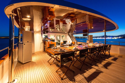 A long dining table set for a meal on a yacht's deck is illuminated against a twilight sky with a distant coastline.