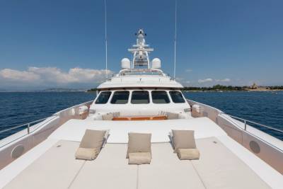 Three lounge chairs with pillows are arranged on the deck of a white yacht, with a clear blue sky and a distant coastline in the background.