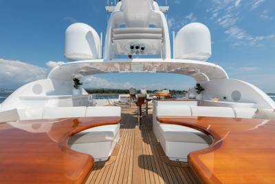 Luxurious white seating and polished wooden tables fill the foreground of a yacht's deck, with a clear blue sky and distant green coastline in the background.