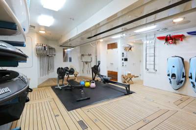 In the foreground, a gym bag and surfboards are visible, while the background features a well-equipped gym with rowing machines, weights, and various exercise equipment.
