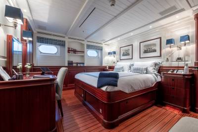 A luxurious yacht cabin features a large bed with blue accents in the foreground, while the background reveals framed artwork and porthole windows.
