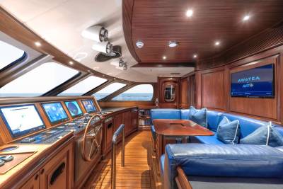 The foreground features a yacht's helm with multiple screens and a wooden steering wheel, while the background reveals a luxurious seating area with blue upholstery and a television screen, all set against a backdrop of the ocean visible through large windows.