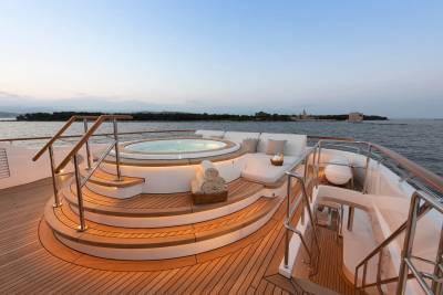 A luxurious yacht's deck features a circular jacuzzi and lounge area in the foreground, with a calm sea and distant coastline in the background.