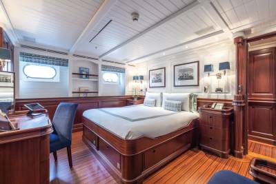 A luxurious yacht cabin features a plush bed in the foreground and a desk with a chair, all set against a backdrop of wood paneling and framed artwork.
