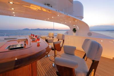 A luxurious yacht deck with a polished wooden bar and plush seating in the foreground overlooks a serene ocean and distant islands at sunset in the background.