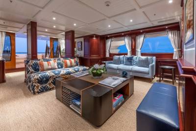 A luxurious yacht salon with a patterned sofa and coffee table in the foreground, and a beach view through open doors in the background.