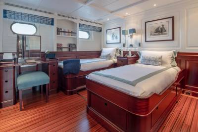 A luxurious yacht cabin features two neatly made beds in the foreground and a vanity desk with a stool and bookshelves in the background.