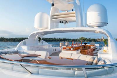 Luxurious outdoor seating and dining areas on a yacht's deck are set against a backdrop of calm water and a distant, tree-lined shore.