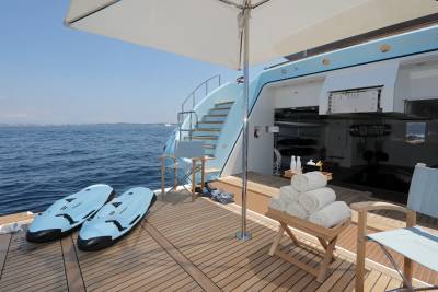 Two blue sea scooters rest on the wooden deck of a yacht in the foreground, while the background reveals a staircase leading up to the yacht's upper deck and the calm blue sea.