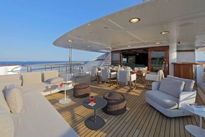 A luxurious yacht deck with comfortable seating in the foreground and a dining area leading into the yacht's interior in the background, all under a clear blue sky.