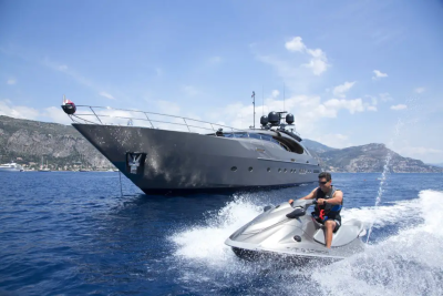 A man on a jet ski speeds across the water in the foreground, with a large, dark yacht anchored behind him and a lush, green coastline in the background.