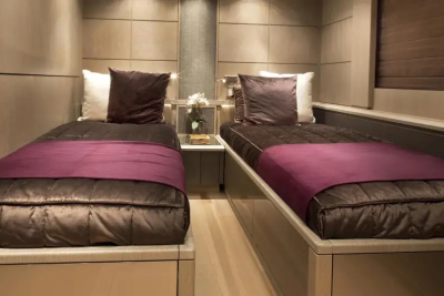 Two neatly made twin beds with plush brown comforters and deep purple throws are positioned in a modern, wood-paneled cabin with soft lighting.