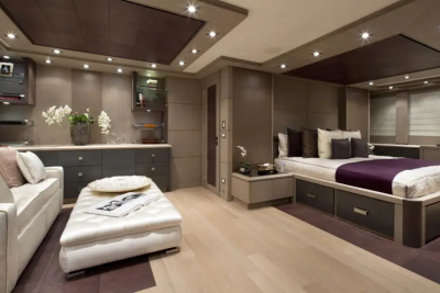A modern yacht cabin features a plush sofa and ottoman in the foreground, with a king-sized bed and built-in cabinetry in the background.