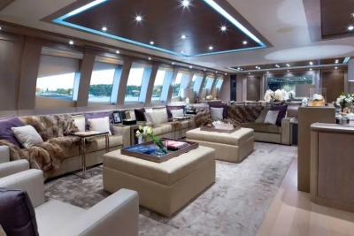 Plush seating and decorative accents fill the foreground of a luxurious yacht lounge, while large windows in the background offer a glimpse of a serene waterfront landscape.