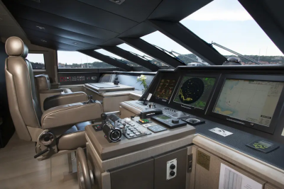 A luxurious yacht's bridge features plush captain's chairs and advanced navigation screens in the foreground, with a blurred view of a sunny harbor and distant hills in the background.