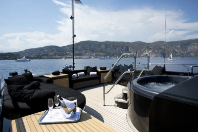 A luxurious yacht deck with a hot tub and seating in the foreground overlooks a calm bay dotted with other boats and a mountainous coastline in the background.