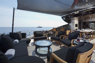Plush black outdoor seating and a dining table are on a yacht's deck, with a calm sea and distant ship in the background.