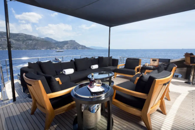A luxurious outdoor seating area with black cushions and wooden chairs on a yacht's deck overlooks a calm blue sea and distant, hazy mountains under a clear sky.
