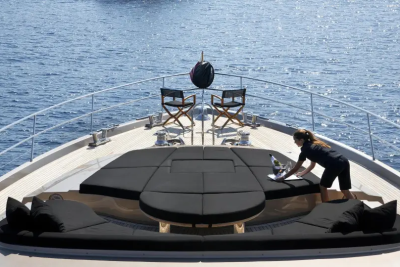 A person cleans a plush, dark seating area on the bow of a yacht, with two director's chairs and the calm blue sea in the background.