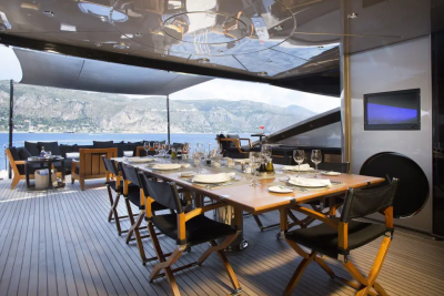 A long dining table is set for a meal on a yacht's deck, with a scenic view of the ocean and hills in the background.