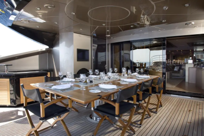 A long wooden dining table set for a meal on a yacht deck is in the foreground, with a glimpse of the ocean and a lounge area visible in the background.