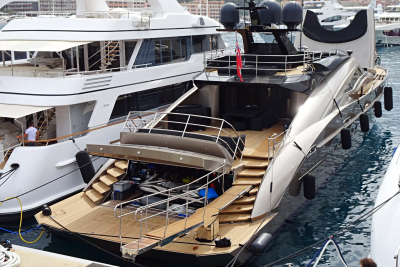 A sleek black yacht with a wooden deck and stairs is docked next to a large white yacht, with other boats and buildings visible in the background.