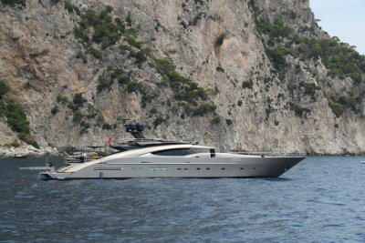 A sleek, modern yacht floats on calm blue water in the foreground, with a towering, rugged cliff face rising steeply behind it.