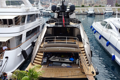 A large, dark yacht with an open stern is docked between two white yachts in a harbor with buildings and hills in the background.
