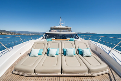 A luxurious yacht with plush lounge chairs in the foreground is anchored in a calm blue sea with a distant coastline under a clear sky.
