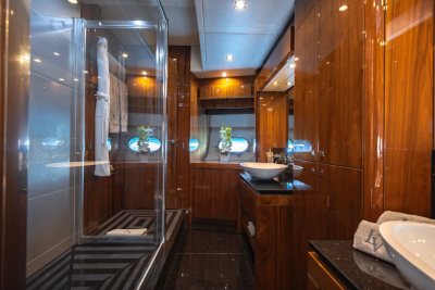 A luxurious yacht bathroom features a glass-enclosed shower in the foreground and a vanity with a sink and a large mirror in the background, all adorned with rich wood paneling.