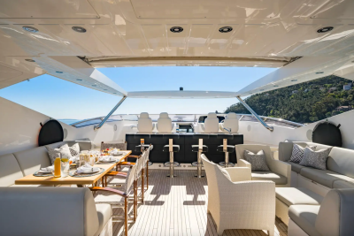 A luxurious yacht deck with a dining table and seating in the foreground, and a bar and helm station with a scenic coastal view in the background.