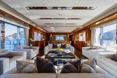 Plush white sofas and a dark wood coffee table fill the foreground of a luxurious yacht's salon, while large windows in the background reveal a sunlit ocean and distant hills.