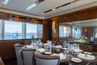 A formal dining table set for a meal is in the foreground, with a large mirror reflecting the room and a view of the ocean visible through windows in the background.