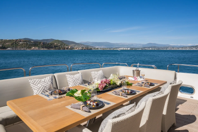 A beautifully set dining table on a yacht's deck overlooks a calm blue sea and a distant, sun-drenched coastline.